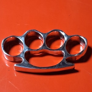 May include: A polished silver knuckle duster with four finger holes and a curved base. The weapon is set against a vibrant orange backdrop.