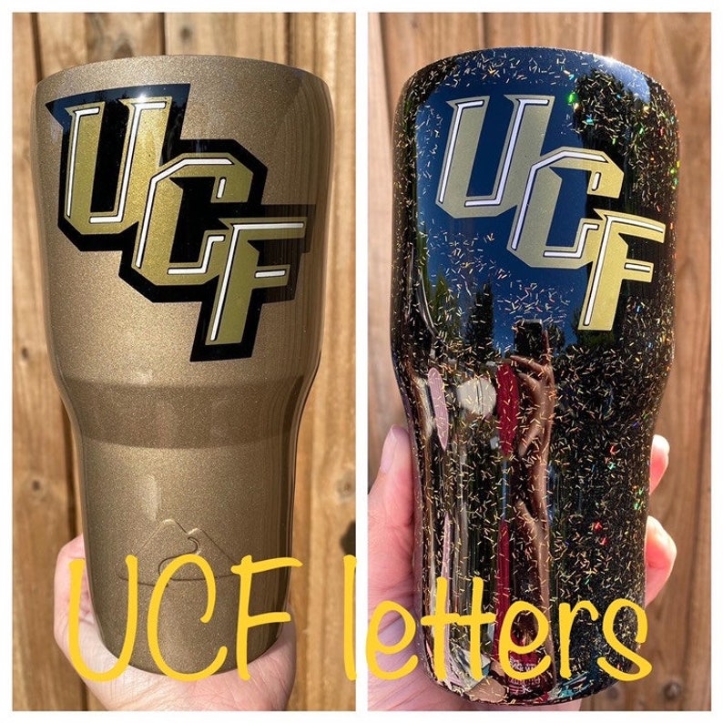 Puede incluir: Dos vasos de acero inoxidable con acabado de purpurina dorada y letras negras que dicen "UCF". El vaso de la izquierda es dorado y el vaso de la derecha es negro.