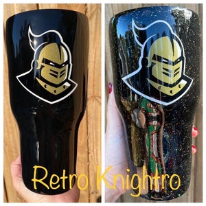 Puede incluir: Un vaso negro y dorado con un logotipo de cabeza de caballero. El vaso tiene un acabado brillante en un lado y un acabado liso en el otro. El texto "Retro Knightro" es visible en la parte inferior de la imagen.