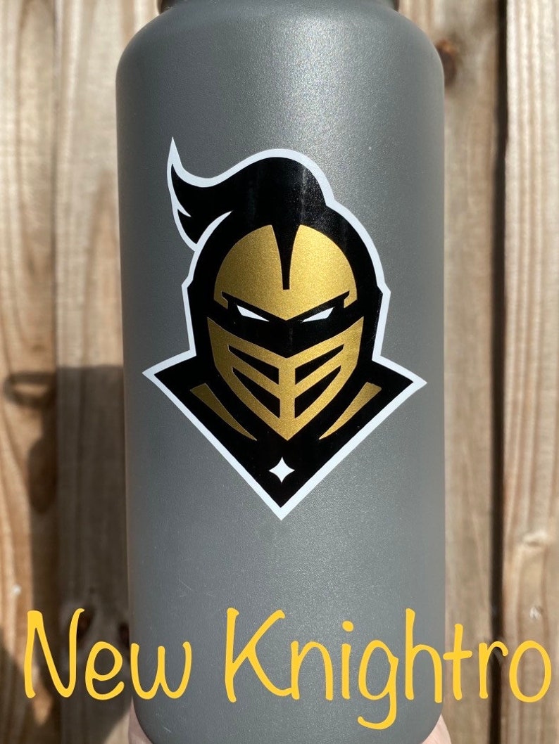 Puede incluir: Una botella de agua gris con una pegatina de logotipo de caballero dorado y negro. La pegatina presenta un casco de caballero con una cara dorada y cabello negro. El texto "New Knightro" es visible en la parte inferior de la imagen.