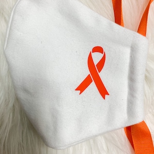 Może przedstawiać: Biała maska ​​na twarz z pomarańczową wstążką. Maska ​​ma dwa pomarańczowe sznurki.