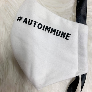 Może przedstawiać: Biała maska ​​na twarz z czarnymi paskami i nadrukiem "#AUTOIMMUNE" z przodu.