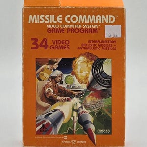 May include: An orange box for the Atari Missile Command video game. The box features the game title, "MISSILE COMMAND," and a graphic of a missile launch. It includes the text "34 VIDEO GAMES" and "INTERPLANETARY BALLISTIC MISSILES + ANTIBALLISTIC MISSILES."