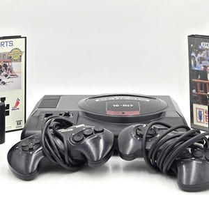 Pode incluir: Console de videogame Sega Genesis de 16 bits preto com dois controladores e vários cartuchos de jogos. Os jogos incluem EA Sports NHL Hockey, Pebble Beach Golf Links, Triple Score e Risk. O console e os acessórios estão em uma superfície branca.