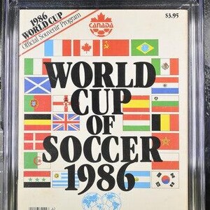 May include: A graded 1986 World Cup Official Souvenir Program. The cover features the text "WORLD CUP OF SOCCER 1986" in large black letters, surrounded by various national flags. The program is encased in a clear protective holder.
