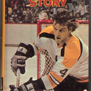 May include: The cover of "The Bobby Orr Story" book shows a hockey player in action, wearing a white, black, and yellow uniform with the number 4. The title is in red and yellow, with the author's name at the bottom.