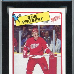 May include: A graded 1988-89 Topps hockey card featuring Bob Probert in a red Detroit Red Wings jersey. The card is graded 8.5 and is labeled as a rookie card. The card is encased in a black holder with the KSA Certification.