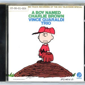 May include: A CD case featuring the artwork from "A Boy Named Charlie Brown." The cover art shows Charlie Brown in a red shirt and cap, holding a baseball mitt. The CD case is in a clear plastic case.