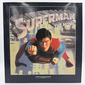 Peut inclure: Une pochette de laserdisc carrée pour Superman The Movie présentant une image centrale d'un personnage volant en costume bleu avec une cape rouge et un emblème sur la poitrine. Le titre Superman The Movie est imprimé en grandes lettres argentées.