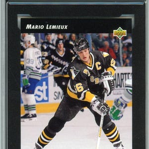 Puede incluir: Una tarjeta de hockey Upper Deck de 1992-93 calificada que presenta a Mario Lemieux con una camiseta negra y amarilla de los Pittsburgh Penguins. La tarjeta está calificada como NMM e incluye el texto "World Junior Grads".