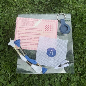 Puede incluir: Un kit de manualidades con una Guía de Puntada Continental, hilo de bordar azul y blanco, un lienzo con una "A" azul y un llavero azul. El kit está empaquetado en una bolsa de plástico transparente, sobre hierba verde.