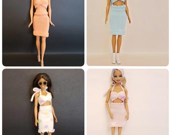 May include: Four dolls are displayed, each wearing a different outfit. The outfits include a peach-colored two-piece set, a light blue two-piece set, a white and polka-dotted skirt and top, and a pink two-piece set. Each doll has different hair and accessories.
