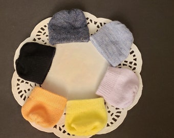 Puede incluir: Seis gorros de bebé de punto de colores diferentes dispuestos en círculo sobre un encaje blanco. Los gorros son en tonos de azul, negro, amarillo, naranja y morado.