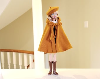 May include: A doll wearing a mustard-colored beret and cape with a black and white striped top and brown boots. The cape has a hood and a tie closure. The doll is standing on a white surface.