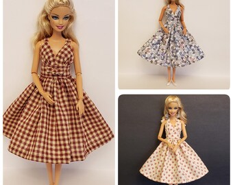 May include: Three Barbie dolls wearing different patterned dresses. The dresses are all sleeveless and have a V-neckline. The dolls are all wearing black shoes.