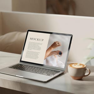 Può includere: Un computer portatile argentato con uno sfondo bianco sullo schermo. Lo schermo mostra la parola "MOCKUP" e del testo sul marchio. Una tazza di caffè con latte art si trova su una superficie bianca accanto al computer portatile.