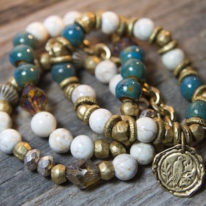 Op de afbeelding: Drie gelaagde armbanden met witte, blauwe en gouden kralen. De armbanden hebben een bedel met een vogelontwerp.