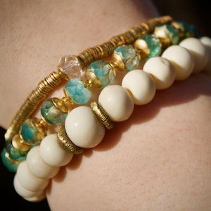 Puede incluir: Un brazalete de capas con cuentas blancas, azules y doradas. El brazalete está hecho de diferentes materiales, incluyendo madera, vidrio y metal.