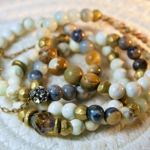 Puede incluir: Un conjunto de cuatro pulseras con cuentas blancas, marrones, azules y doradas. Las pulseras están superpuestas y tienen una variedad de texturas y formas.