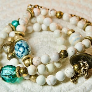 Könnte beinhalten: Ein weißes und türkisfarbenes Perlenarmband mit goldenen Akzenten. Das Armband hat eine blau-weiße Perle mit Muster, eine türkisfarbene Glasperle und einen goldenen Knopf mit der Gravur "Ruby".