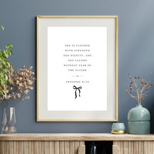May include: A framed print with a white background and black text that reads "She is clothed with strength and dignity, and she laughs without fear of the future. Proverbs 31:25". A black bow is at the bottom of the text.