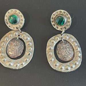 Könnte beinhalten: Ein Paar silberne Ohrringe mit einer grünen Abalone-Muschel und einem silbernen Gesichtsdesign. Die Ohrringe sind oval geformt und haben eine strukturierte Oberfläche.