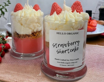 May include: Two glass jar candles resembling strawberry shortcake. Each candle has a red base, a layer of crumbs, white frosting, and strawberry-shaped wax pieces. The label reads "HELLO ORGANIC Strawberry Shortcake". Each candle is 12 ounces / 340 grams.
