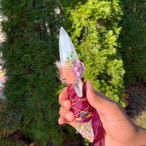 Könnte beinhalten: Ein klarer, spitzer Kristallstab, verziert mit getrockneten Blumen, Federn und einem bordeauxfarbenen Band. Der Kristall ist mit bordeauxfarbenem Stoff und goldfarbenem Garn umwickelt. Der Stab wird vor einem Hintergrund aus grünem Laub gehalten.