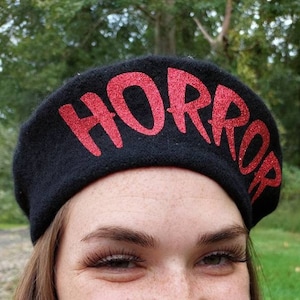 K&ouml;nnte beinhalten: Eine schwarze Baskenm&uuml;tze mit dem Wort "HORROR" in roten Glitzer geschrieben.