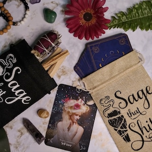 Könnte beinhalten: Zwei kleine Jutesäcke mit dem Aufdruck "Sage that Shit". Die Säcke sind mit Salbeibündeln gefüllt und liegen auf einer weißen Oberfläche. Auf dem Bild sind auch einige Kristalle, ein Tarotkartenspiel und ein Salbei-Reinigungsstab zu sehen.