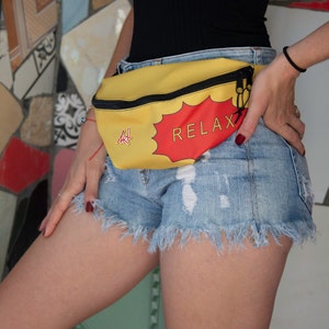 May include: A yellow fanny pack with a red speech bubble that says "RELAX" in black text. The fanny pack has a black zipper and is worn around the waist of a person wearing denim shorts.