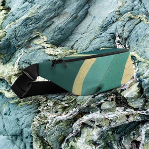 Puede incluir: Una riñonera verde azulado con una correa y hebilla negras ajustables. La bolsa presenta una cremallera dorada y un diseño de rayas diagonales en verde azulado, dorado y patrones texturizados. La riñonera está diseñada para usarse alrededor de la cintura.