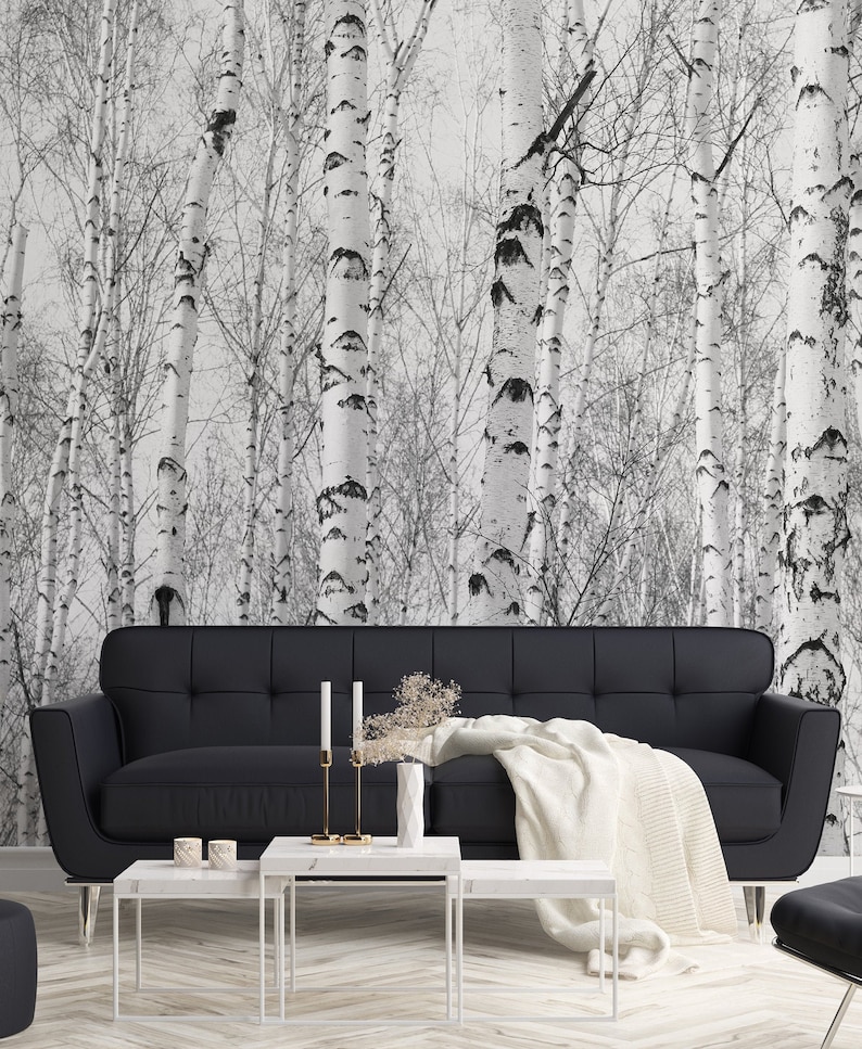 May include: A black leather sofa with a white throw blanket sits in front of a wall with a black and white birch tree wallpaper. Three white marble coffee tables are in front of the sofa.