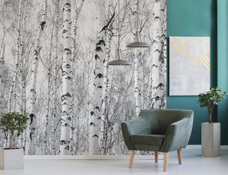 May include: A green armchair with wooden legs sits in front of a wall with a black and white birch tree wallpaper. A teal wall is to the right of the armchair with a gray canvas with gold lines hanging on it. Two pendant lights hang from the ceiling above the armchair.