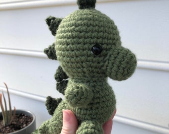 May include: A green crocheted dinosaur toy with black eyes. The dinosaur has a spiky back and a long tail.