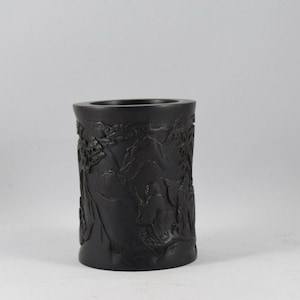 Puede incluir: Un portapinceles cilíndrico negro con intrincados tallados de una escena de montaña y figuras. Los tallados son detallados y muestran un alto nivel de artesanía.