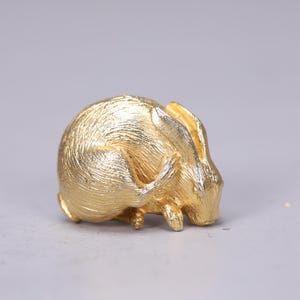 May include: A gold-toned metal figurine shaped like a rabbit in a curled, resting position. The surface of the rabbit features a textured, brushed finish that mimics the appearance of fur, with detailed ears and a rounded body form.