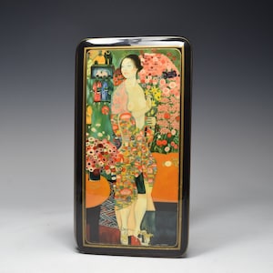 Puede incluir: Una caja de madera lacada en negro con una pintura colorida de una mujer de pie en un jardín con flores. La mujer lleva un vestido largo estampado y tiene el pelo largo y oscuro. La caja es rectangular y tiene una tapa con bisagras.