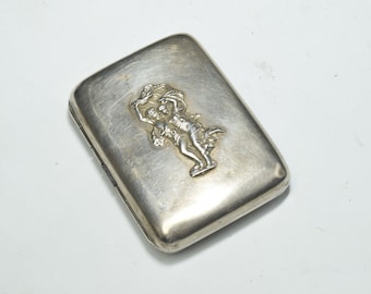 May include: A silver cigarette case with an embossed design of two figures holding a wreath.