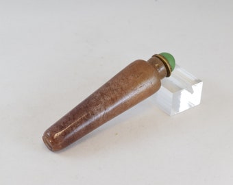 May include: A small, brown, cylindrical bottle with a green glass stopper. The bottle is made of a smooth, polished material and has a slightly tapered shape.