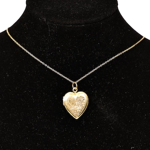 Puede incluir: Un collar con un colgante en forma de corazón dorado. El relicario cuelga de una delicada cadena y presenta un diseño floral grabado. El collar se muestra sobre un fondo negro.