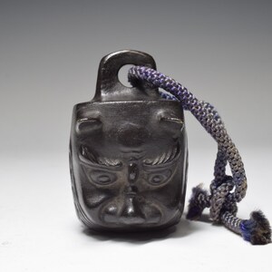 May include: A small, black, metal bell with a demon face design. The bell has a dark blue cord attached to it.