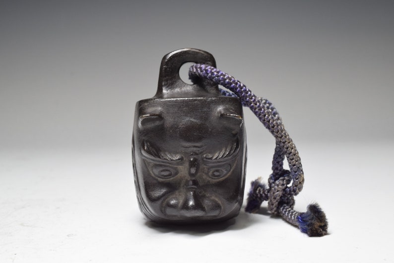 May include: A small, black, metal bell with a demon face design. The bell has a dark blue cord attached to it.
