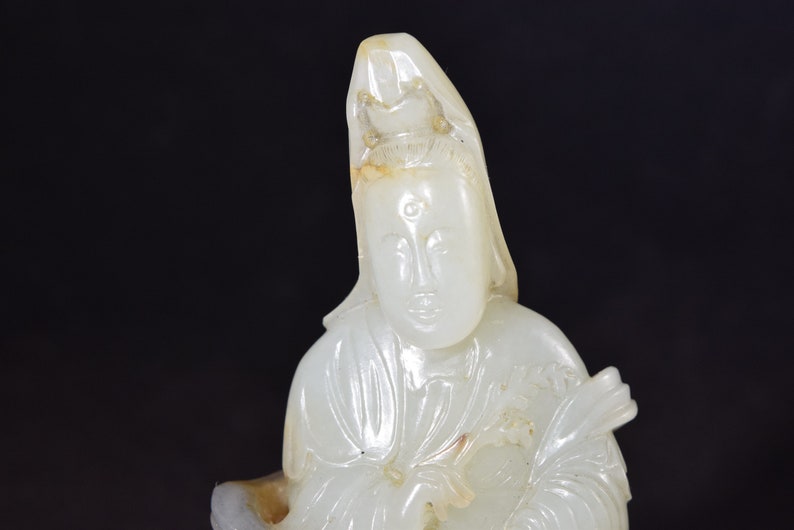 Puede incluir: Una escultura de jade blanco de una figura sentada con una corona y un tercer ojo en la frente. La figura lleva una t&uacute;nica larga y tiene las manos juntas delante de ella.
