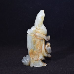 Puede incluir: Una escultura de jade blanco de una mujer con una t&uacute;nica fluida, sosteniendo un p&aacute;jaro en su mano. La escultura est&aacute; tallada con intrincados detalles y tiene un tono c&aacute;lido y terroso.
