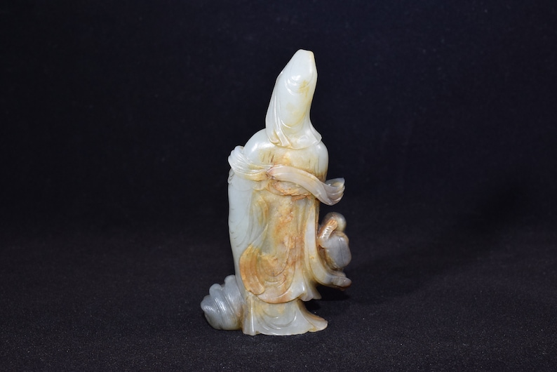 Puede incluir: Una escultura de jade blanco de una mujer con una t&uacute;nica fluida, sosteniendo un p&aacute;jaro en su mano. La escultura est&aacute; tallada con intrincados detalles y tiene un tono c&aacute;lido y terroso.