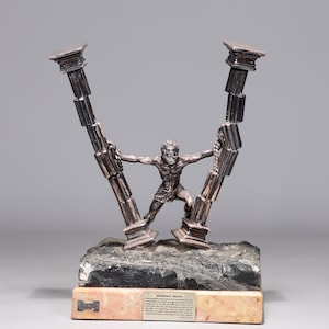 Puede incluir: Una escultura de bronce de un hombre sosteniendo dos pilares. La escultura está sobre una base de mármol negro y rosa. La base tiene una placa que dice "La muerte de Sansón".