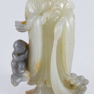 Puede incluir: Una escultura de jade blanco de una figura con ropas fluidas y un ni&ntilde;o a su lado. La escultura est&aacute; tallada con un estilo detallado e intrincado.