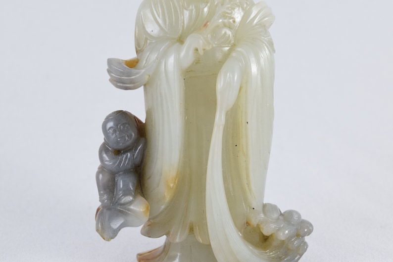 Puede incluir: Una escultura de jade blanco de una figura con ropas fluidas y un ni&ntilde;o a su lado. La escultura est&aacute; tallada con un estilo detallado e intrincado.