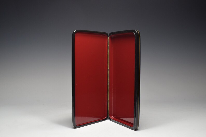 May include: A black lacquered box with a red interior. The box is open and the interior is visible. The box is made of wood and has a gold trim.